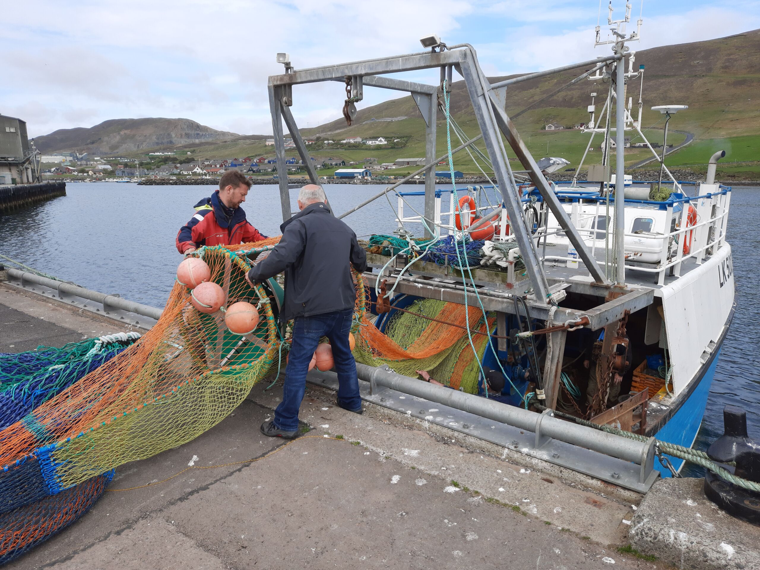 two white men stand at a dock load a fishing net into a medium-sized trawling vessel