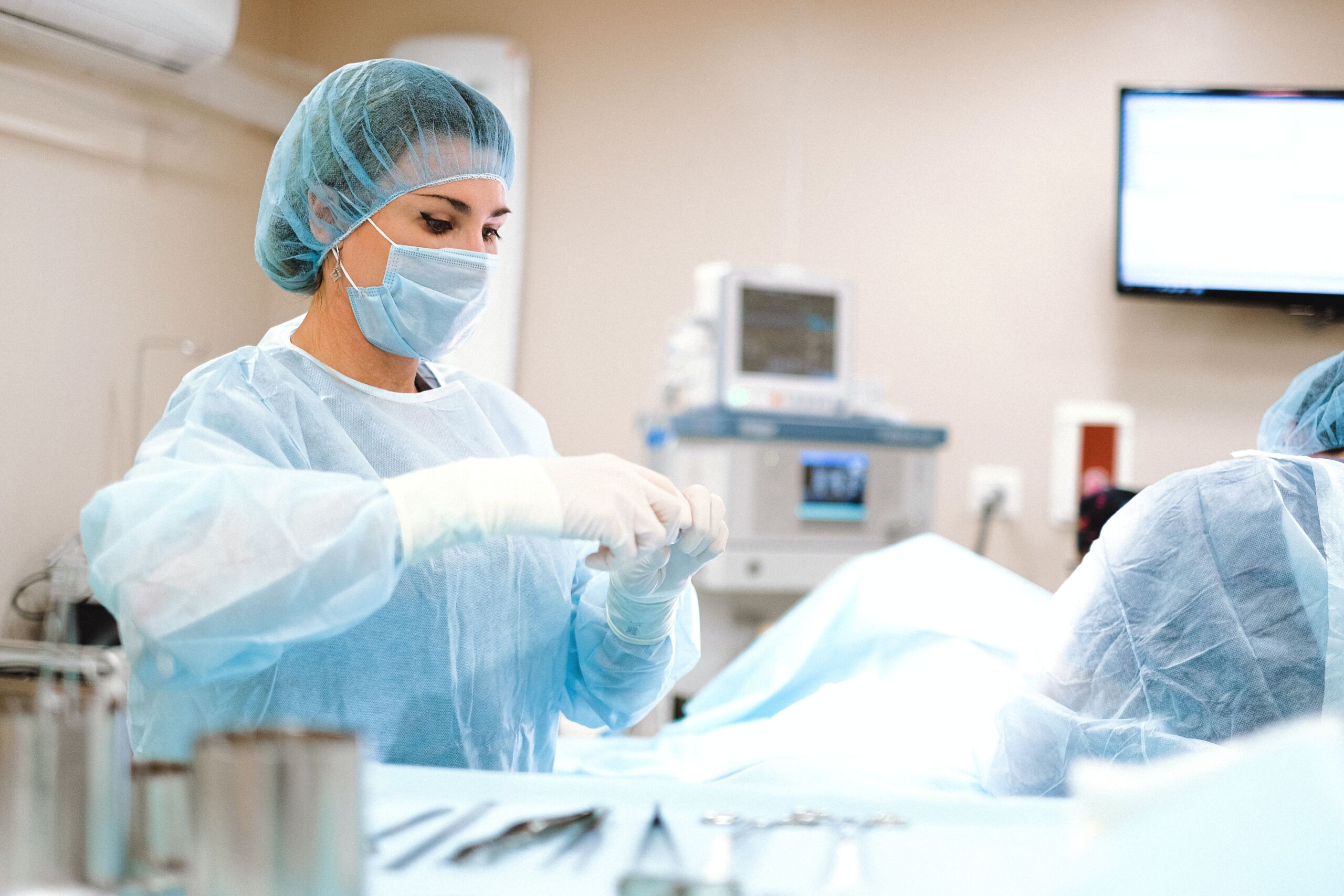 A woman in a surgical mask and gown is standing over a hospital bed with a surgical tool in her gloved hands