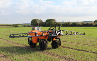 A small orange vehicle on 4 wheels moves across a field. It has a light metal glider on top that stretches out on either side.